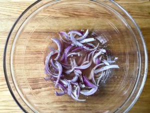 Sliced red onions in bowl with oil, vinegar, salt, and pepper.