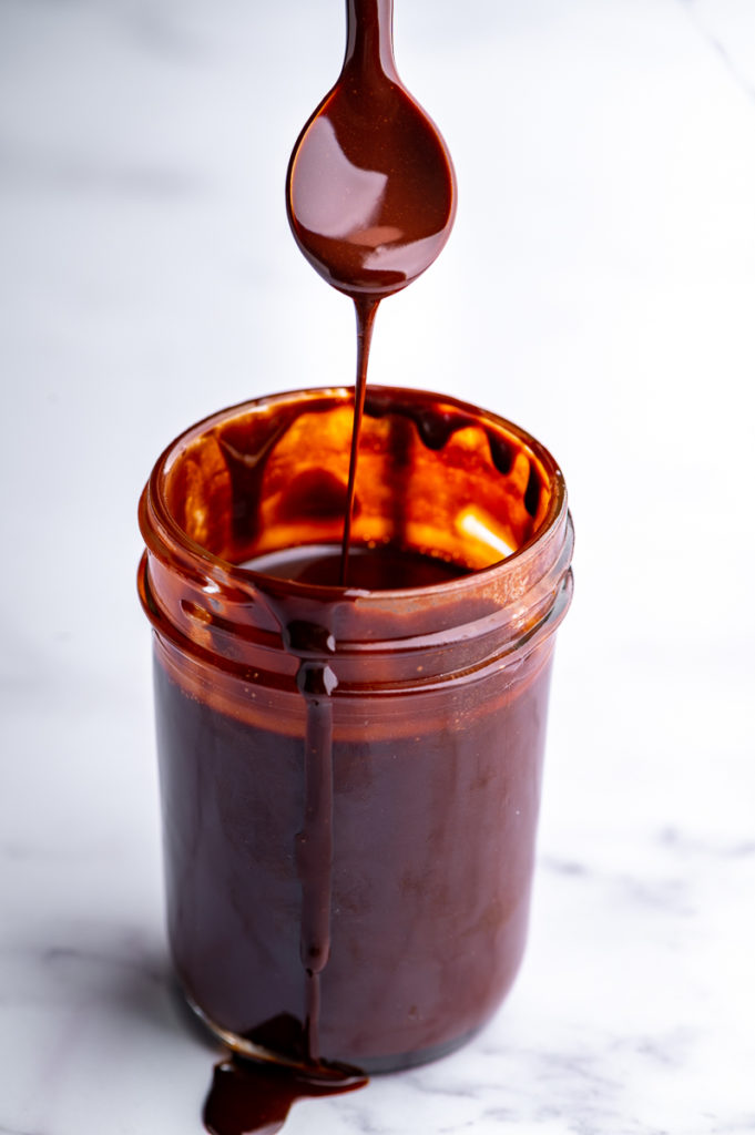 A spoon dripping chocolate syrup lifted from a jar of chocolate syrup.
