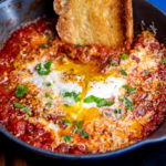 Eggs in purgatory in a small skillet with with a hunk of crusty bread dipping into the sauce.