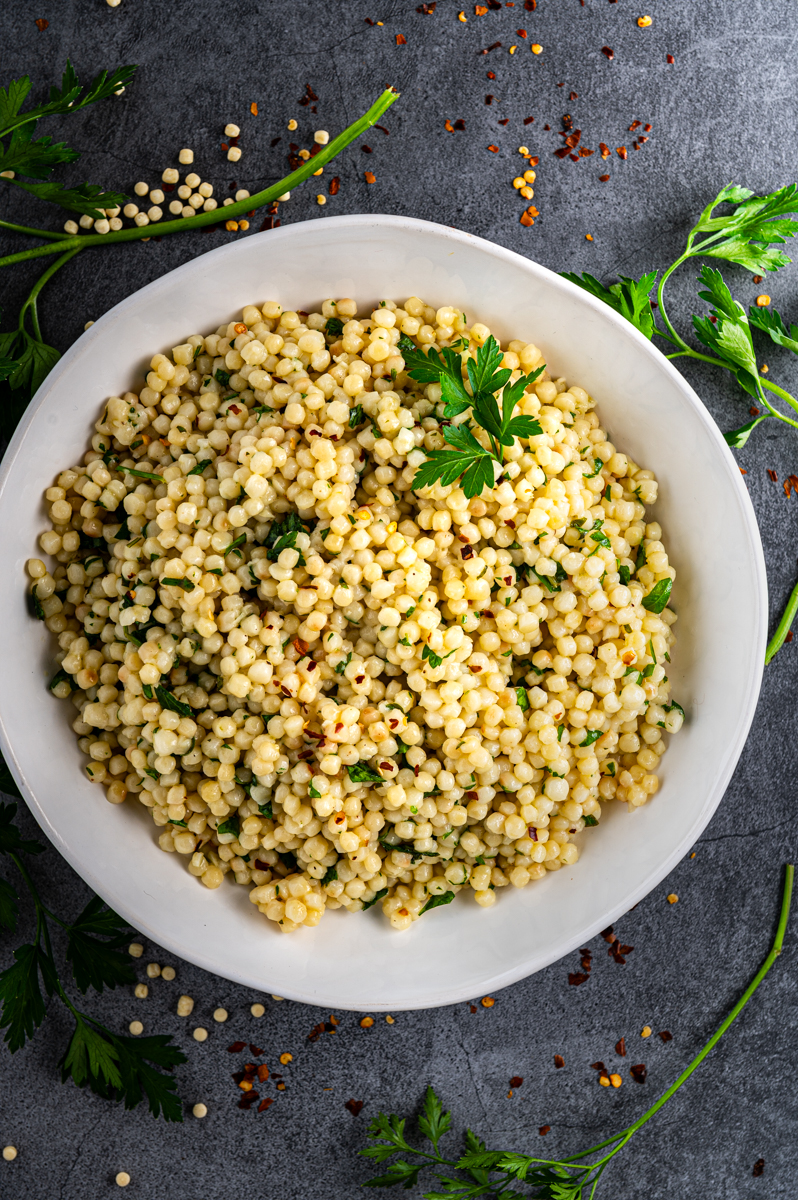 A white bowl filled with lemon herb pearl couscous with parsley sprigs surrounding bowl.