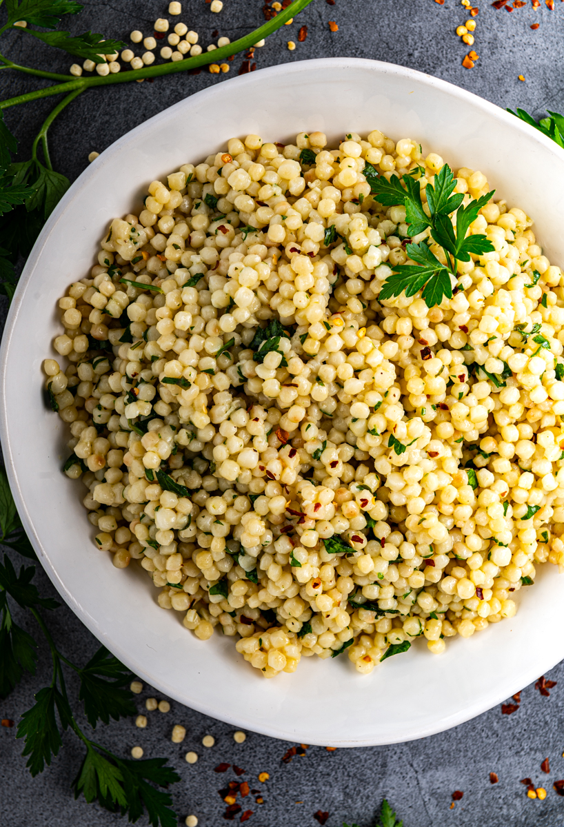 A white bowl filled with lemon herb pearl couscous with parsley sprigs surrounding bowl.