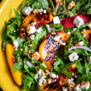 Grilled peaches and arugula salad tossed in balsamic vinaigrette in a yellow bowl.