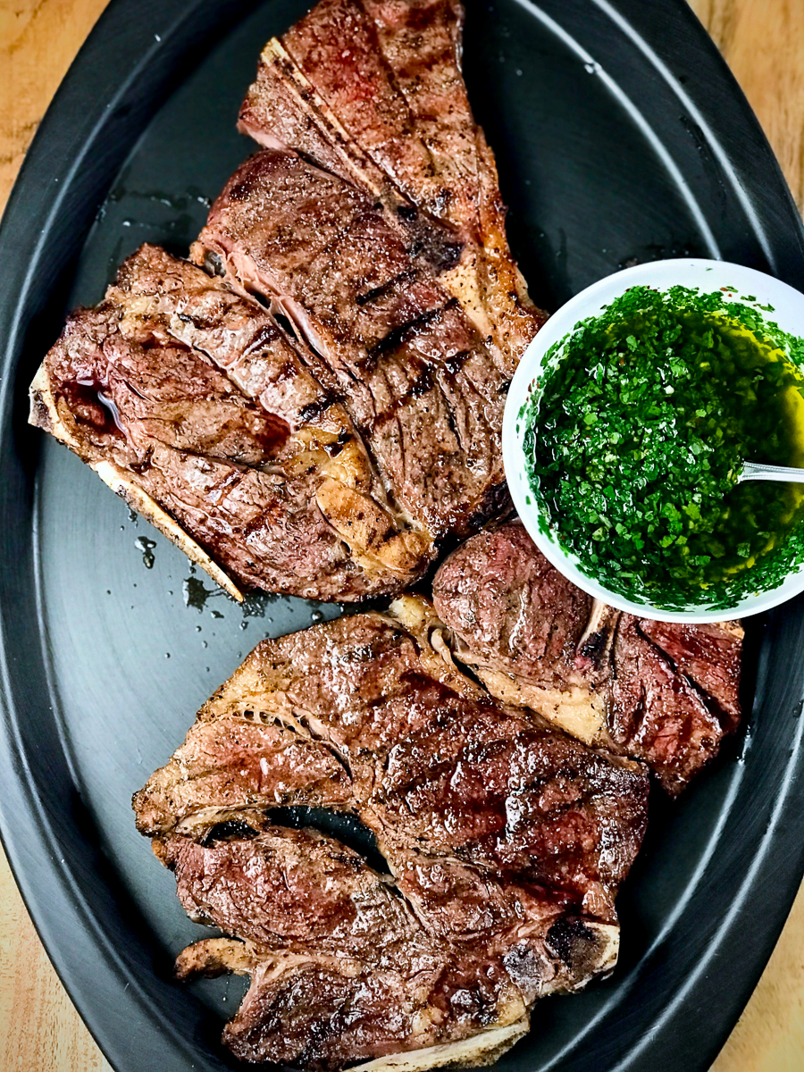 Grilled 7-bone chuck steaks on a platter with a bowl of chimichurri.