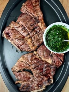 Grilled 7-bone chuck steaks on a platter with a bowl of chimichurri.