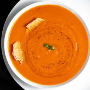 A white bowl of creamy tomato soup with 2 pieces of frico cheese.