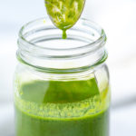 A jar of basil vinaigrette with a spoon over it and a drop of vinaigrette falling back into the jar.