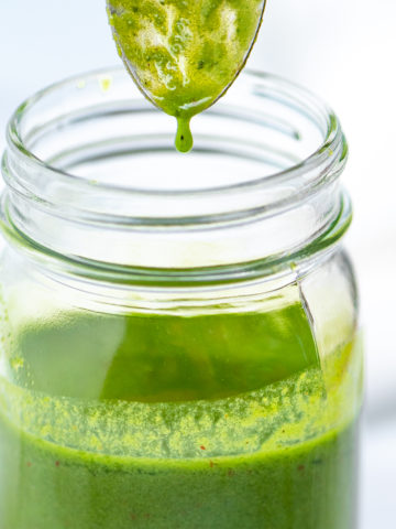 A jar of basil vinaigrette with a spoon over it and a drop of vinaigrette falling back into the jar.