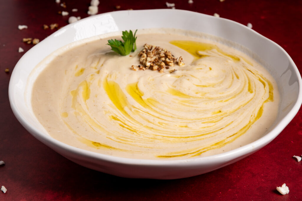 A bowl of cauliflower soup in a white bowl on a dark red board drizzled with olive oil.