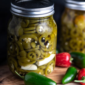 Two jars of pickled jalapeños with scattered jalapeño peppers on a board.