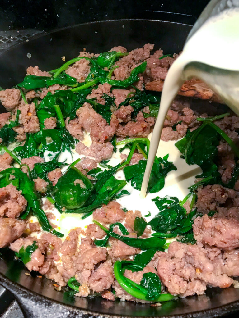 Pouring heavy cream into skillet with browned sausages and wilted spinach.