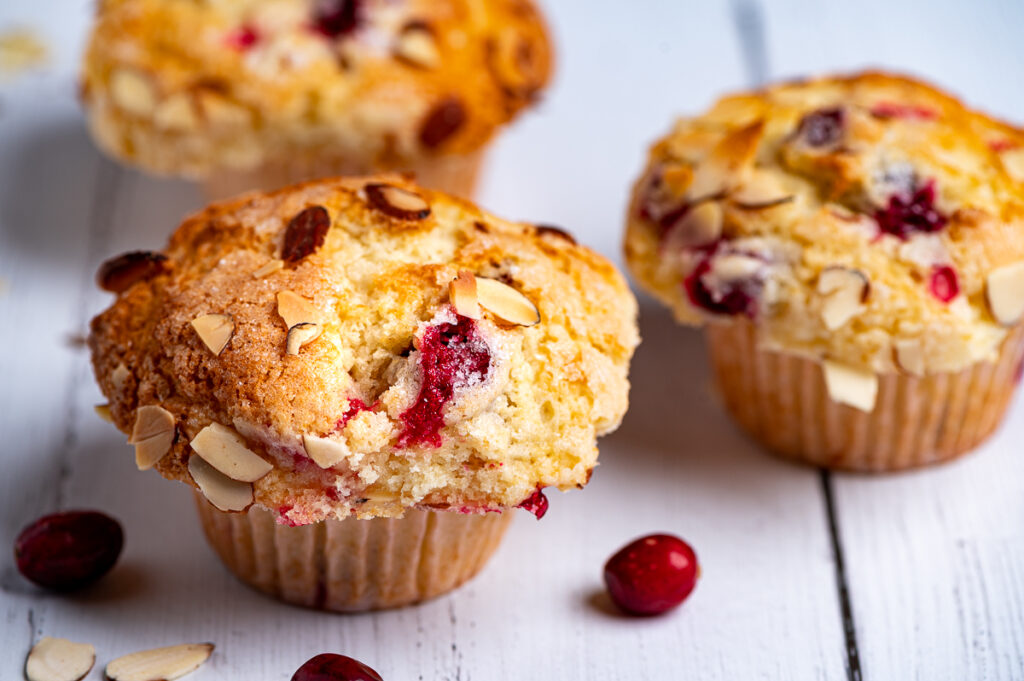 Three cranberry muffins on a board with scattered cranberries.
