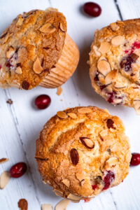 Three cranberry muffins on a board with scattered cranberries.