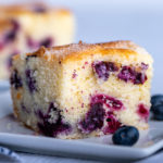 A slice of blueberry cake with a few blueberries surrounding it.