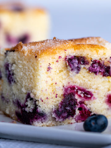 A slice of blueberry cake with a few blueberries surrounding it.
