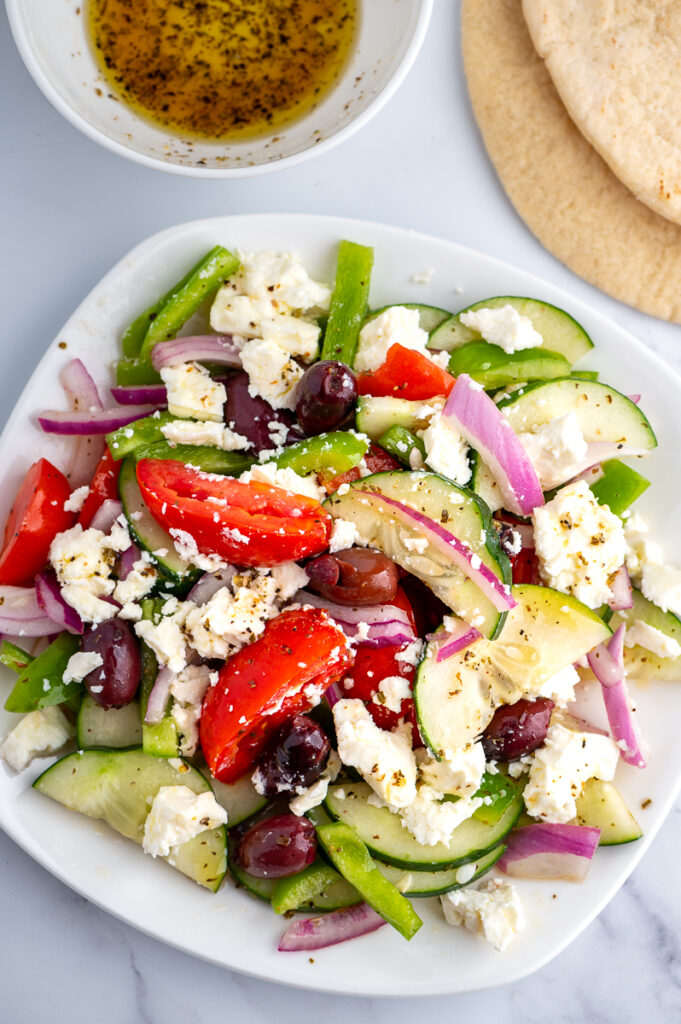 Greek salad on a white plate with dressing in a bowl and a slice of pita.
