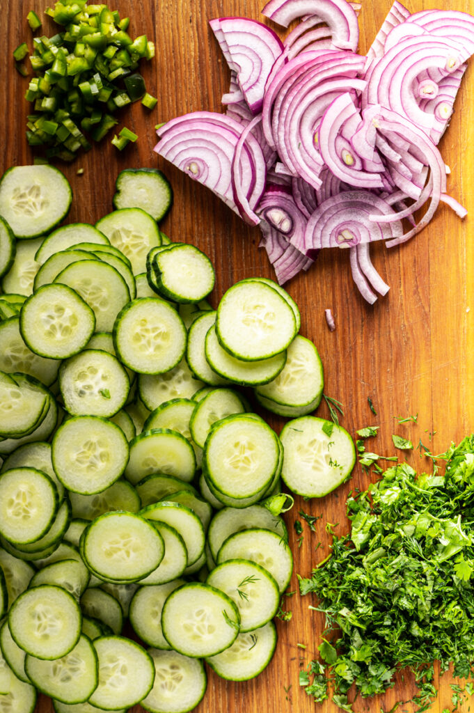 Sliced cucumbers, red onions and chopped herbs on a cutting board.